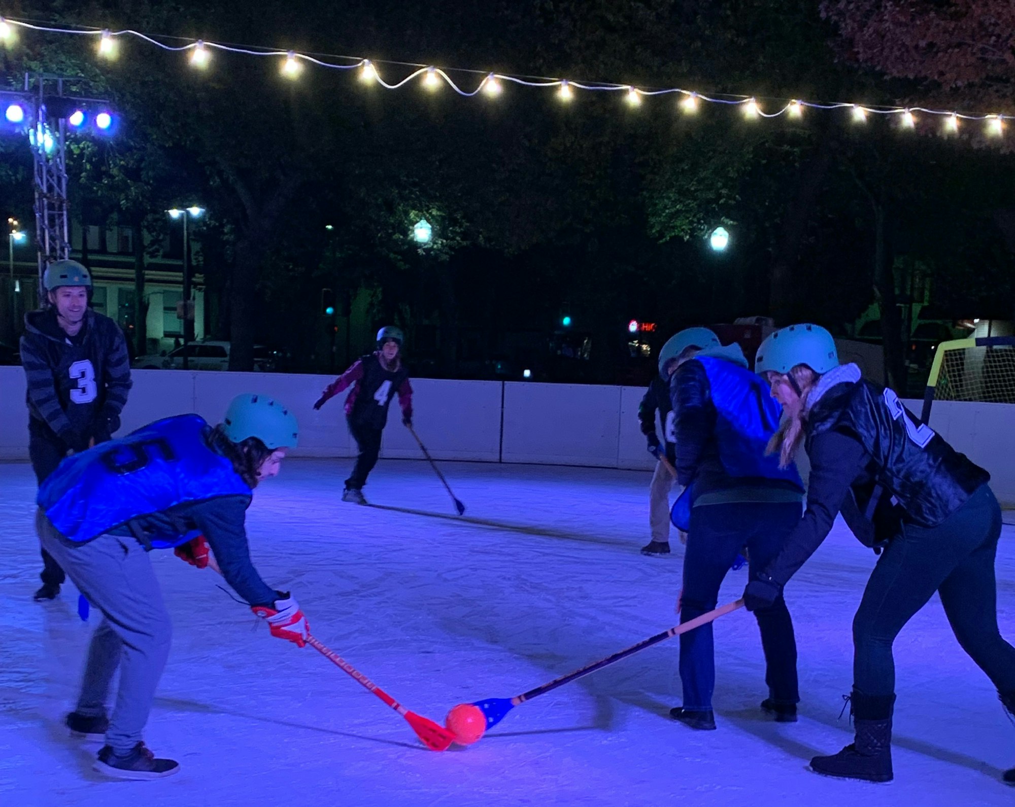 People playing broomball on an ice rink at night, wearing helmets, under string lights.