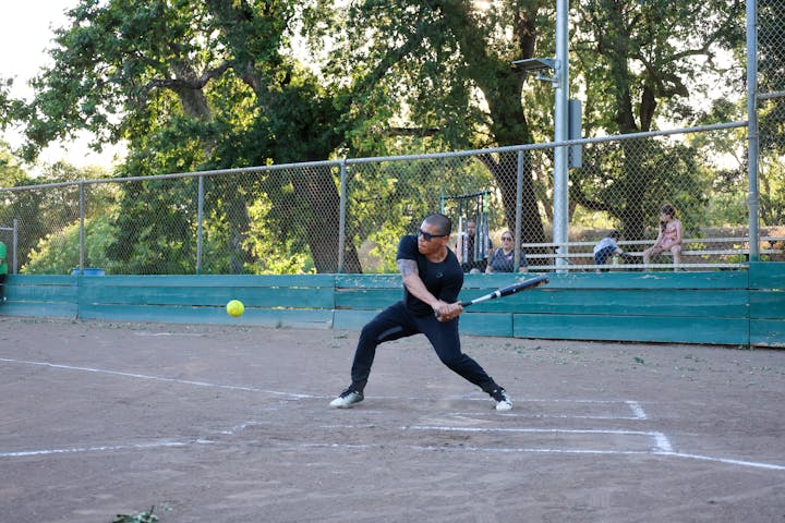 A person is playing softball, about to hit a pitched ball, with spectators in the background.