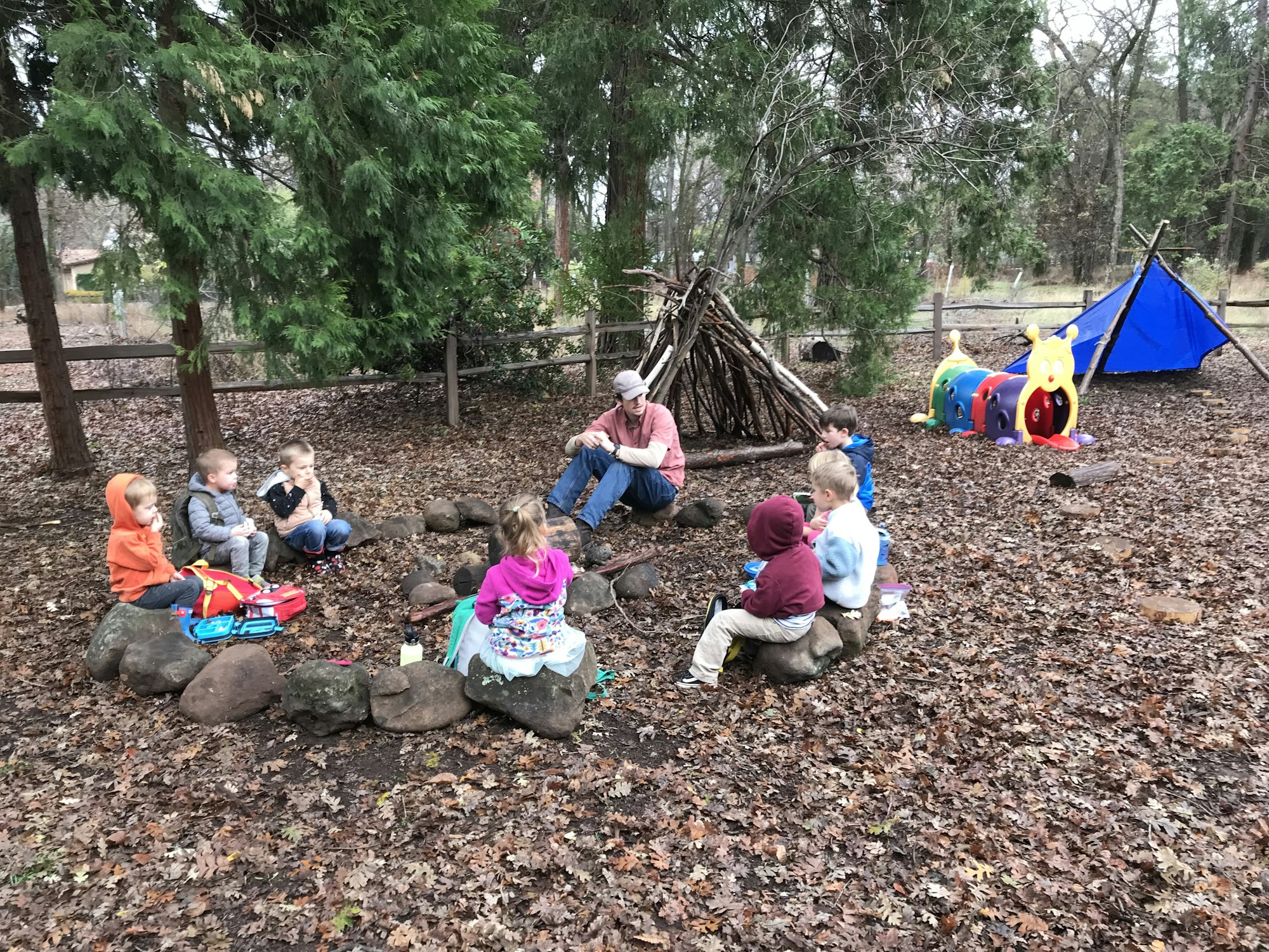 Children and an adult sit in a circle on rocks in a wooded area, with a stick shelter and colorful play tunnel nearby.