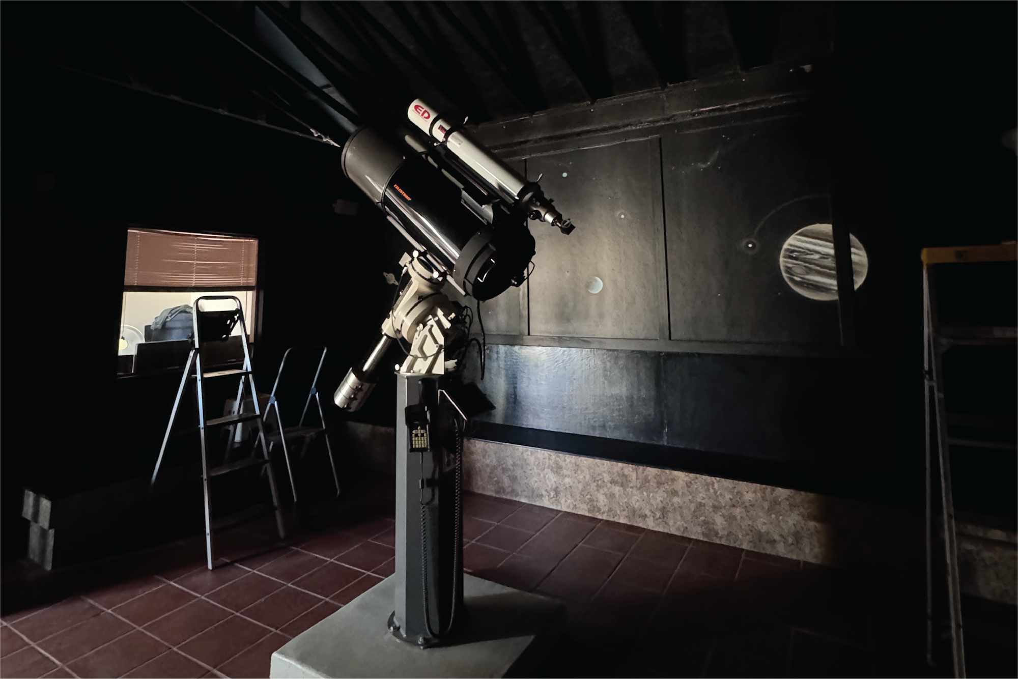 A telescope on a mount in a dark room with a window and a ladder nearby.