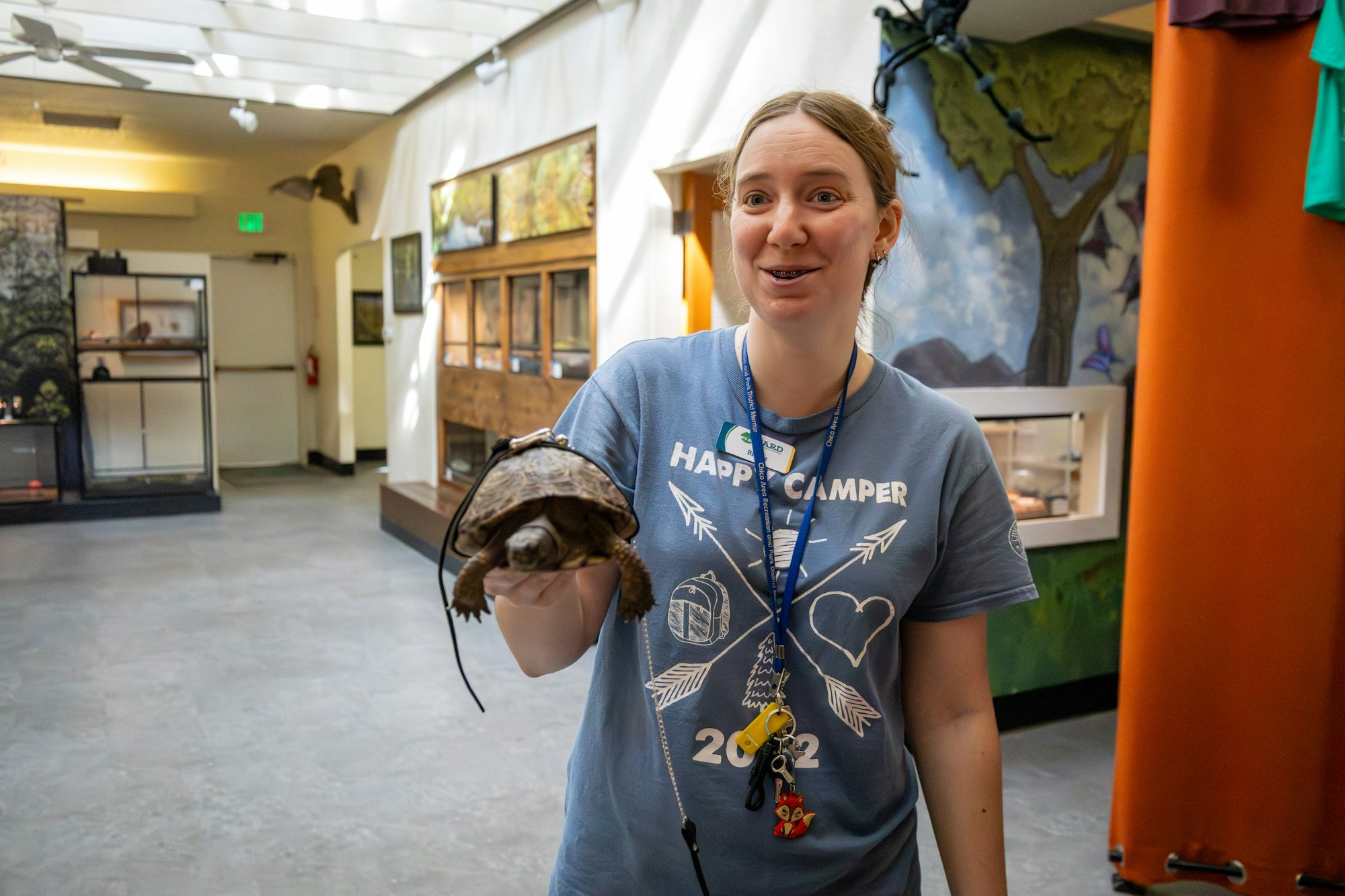 A person in a blue shirt holds a turtle, smiling in a bright indoor space with animal exhibits in the background.