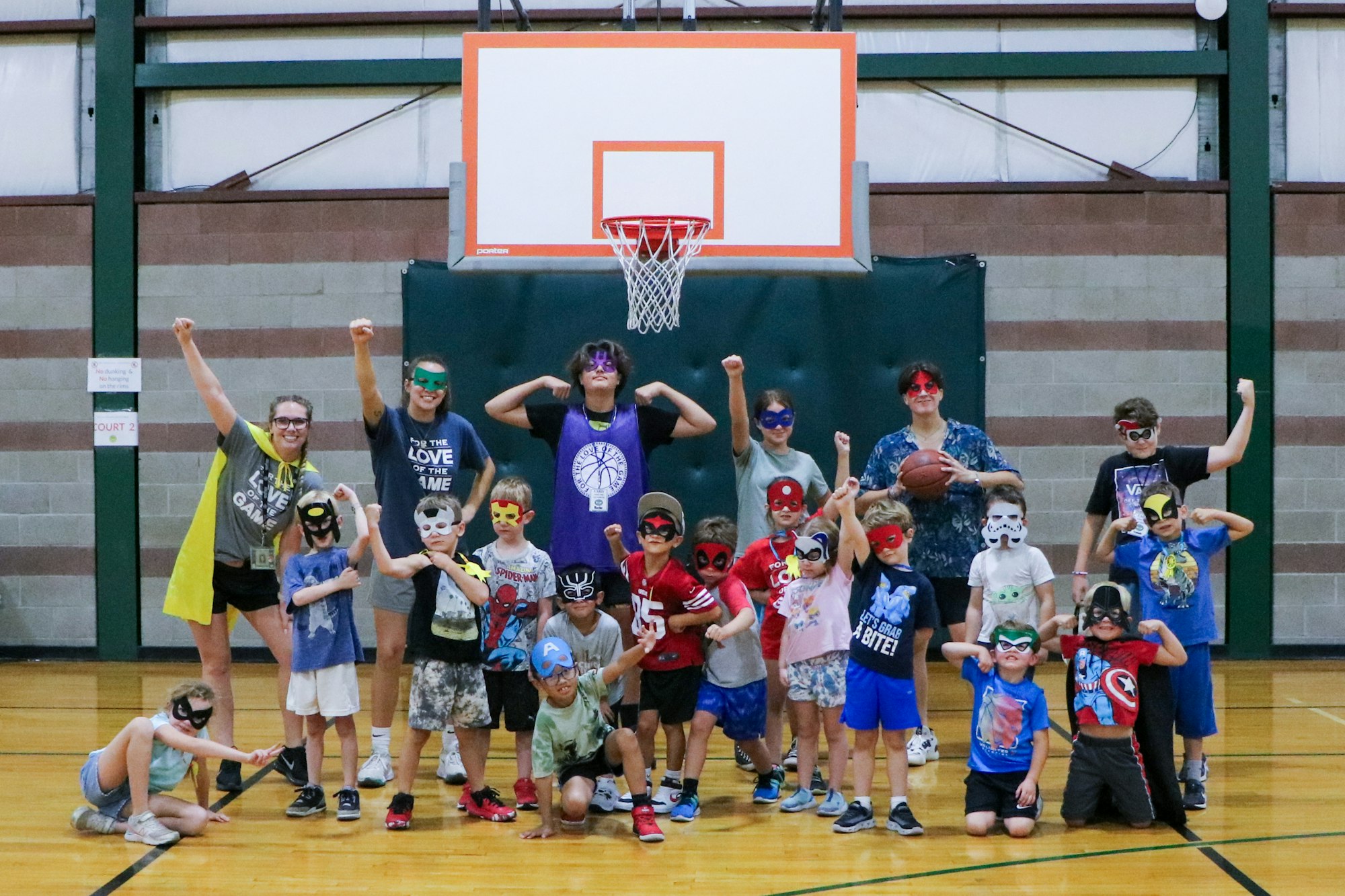 Group of children and adults in masks posing in a gym with a basketball hoop in the background.