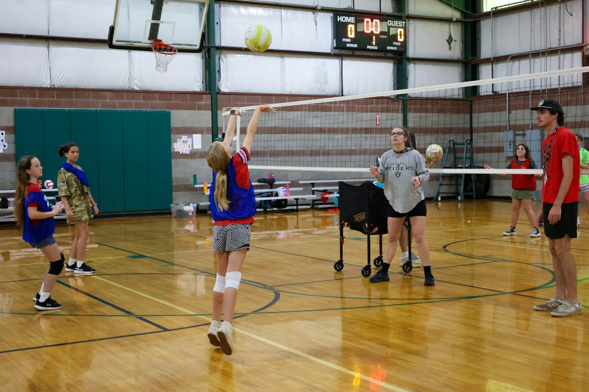 A group of people playing volleyball in an indoor gym, with one person jumping near the net.