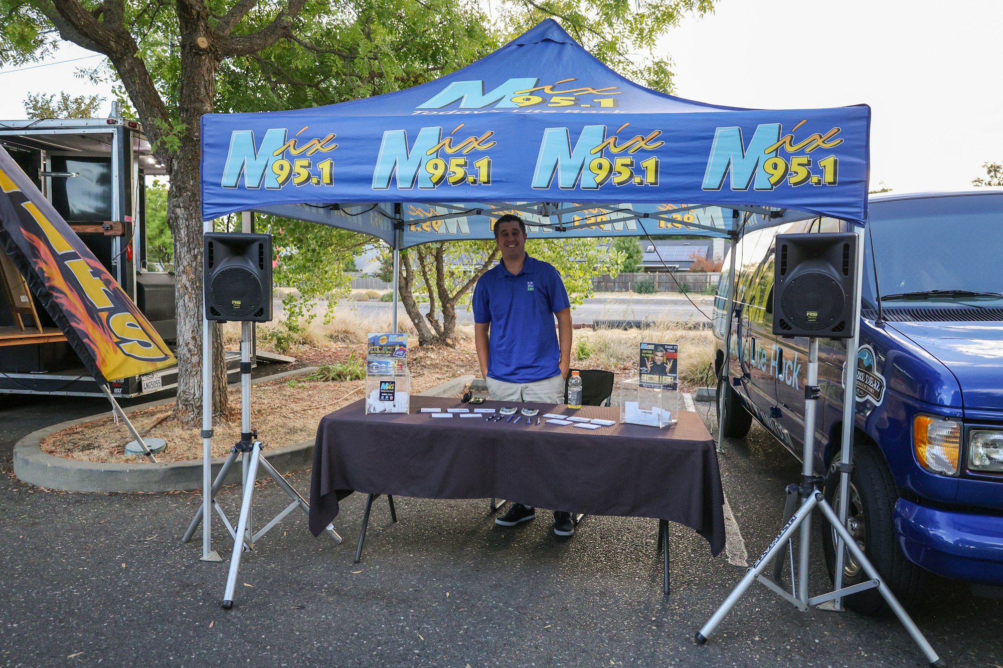 A promotional booth with a blue canopy for "Mix 95.1," featuring a man standing at a table with flyers and speakers nearby.