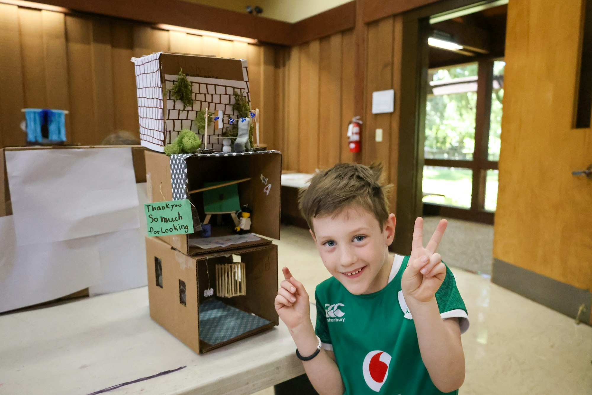 A child showing a cardboard house model with a "Thank you so much for looking" sign, smiling and making peace signs.