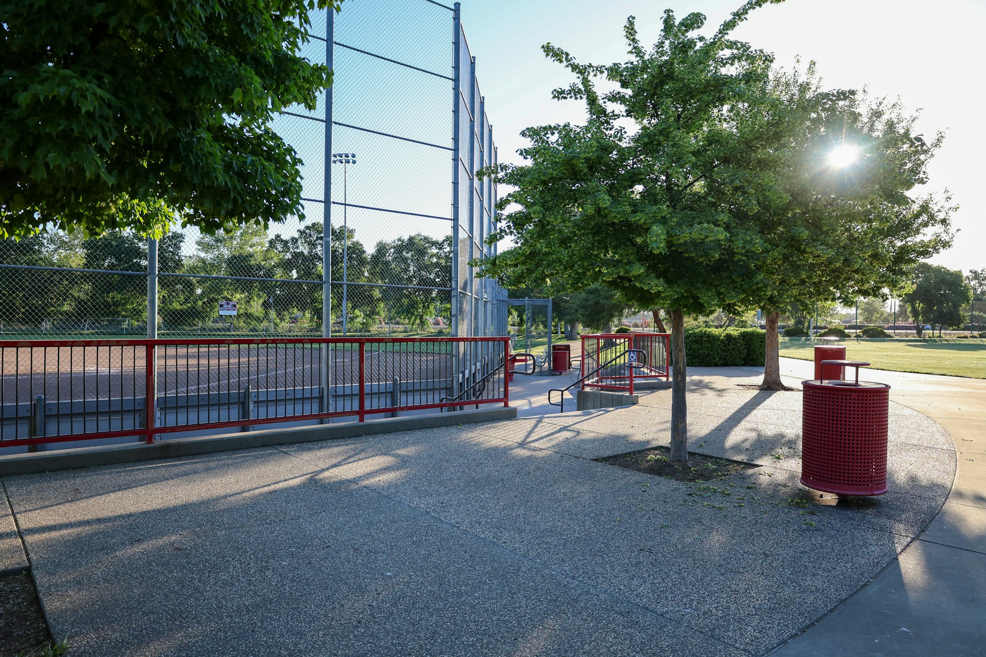 Public park with a baseball diamond, trees, benches, and a trash can on a sunny day.