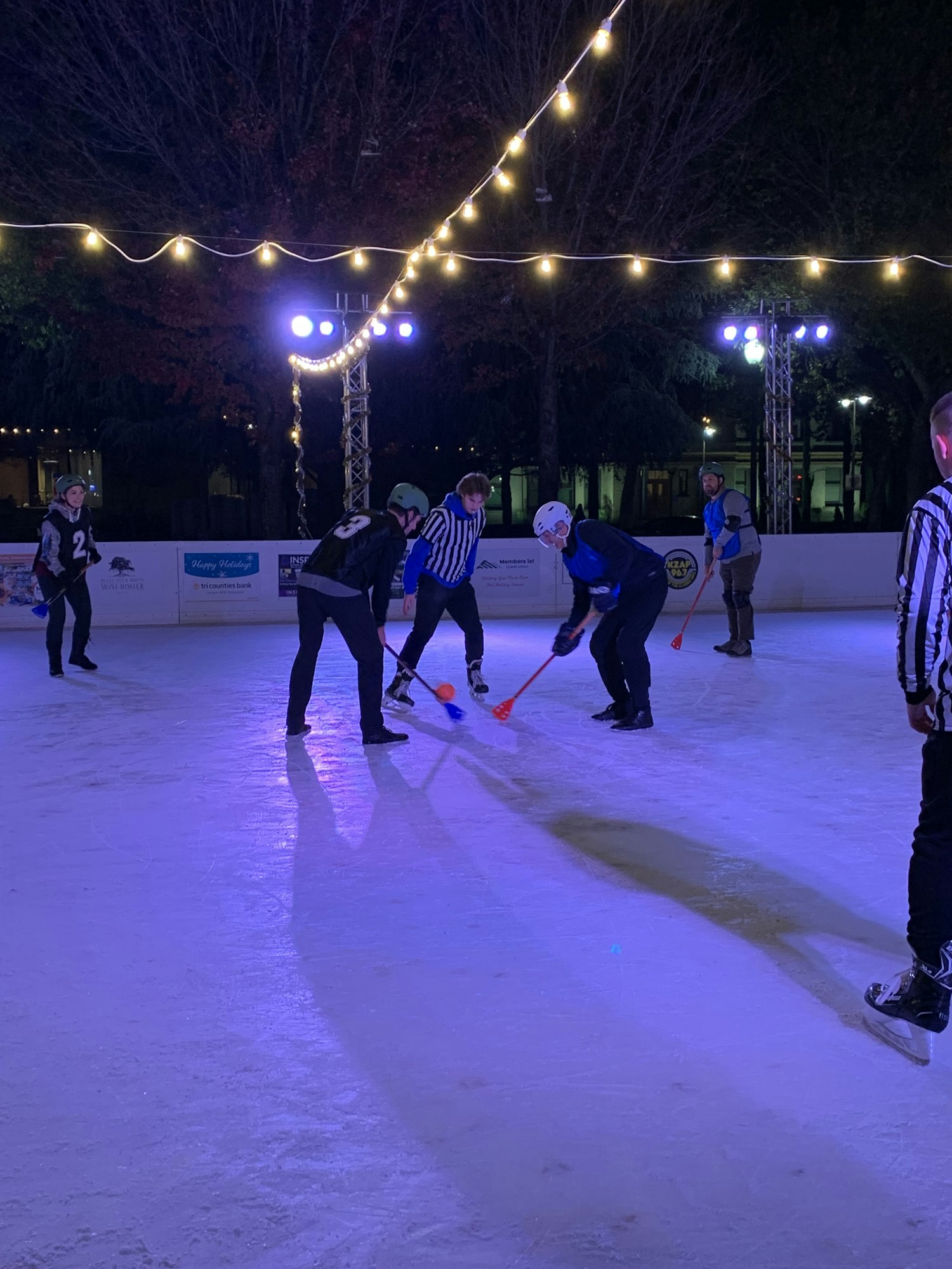 People are playing broomball on an ice rink at night under string lights.