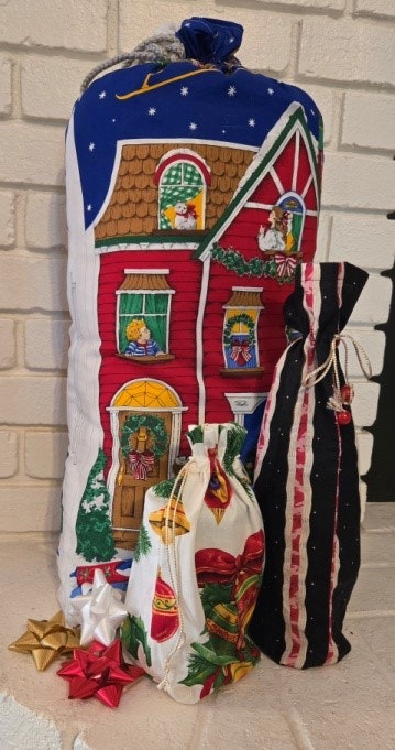 Three decorative fabric bags with Christmas designs, including a house motif, next to gold and red bows on a white brick background.