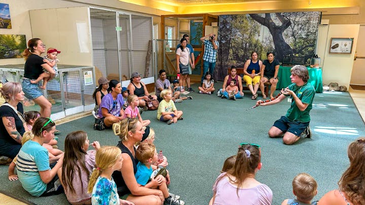 An educator with a snake presenting to an attentive audience of children and adults indoors.