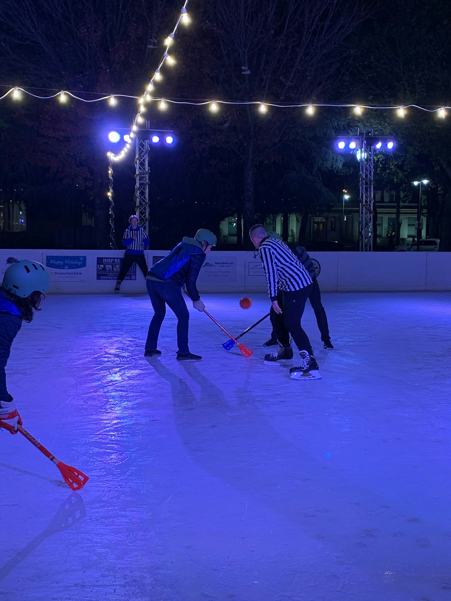 People playing a game on an ice rink under colorful lights, holding sticks and wearing helmets.