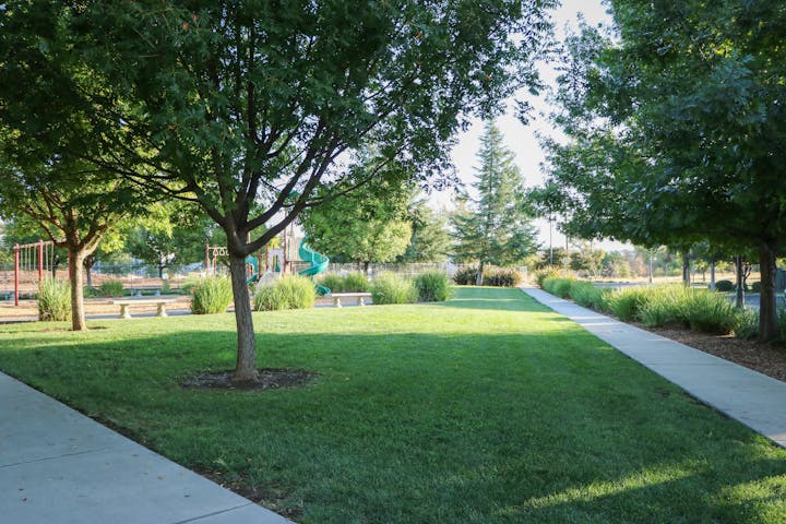 A well-maintained park with green grass, trees, a playground with a slide, and walking paths.