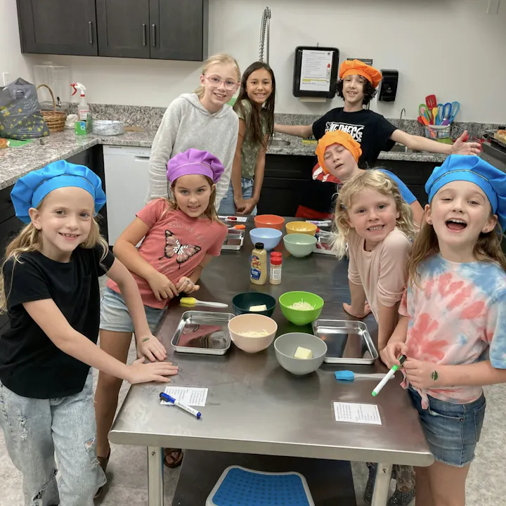 A group of children in colorful chef hats are gathered around a kitchen counter with bowls and cooking ingredients, smiling and engaging.
