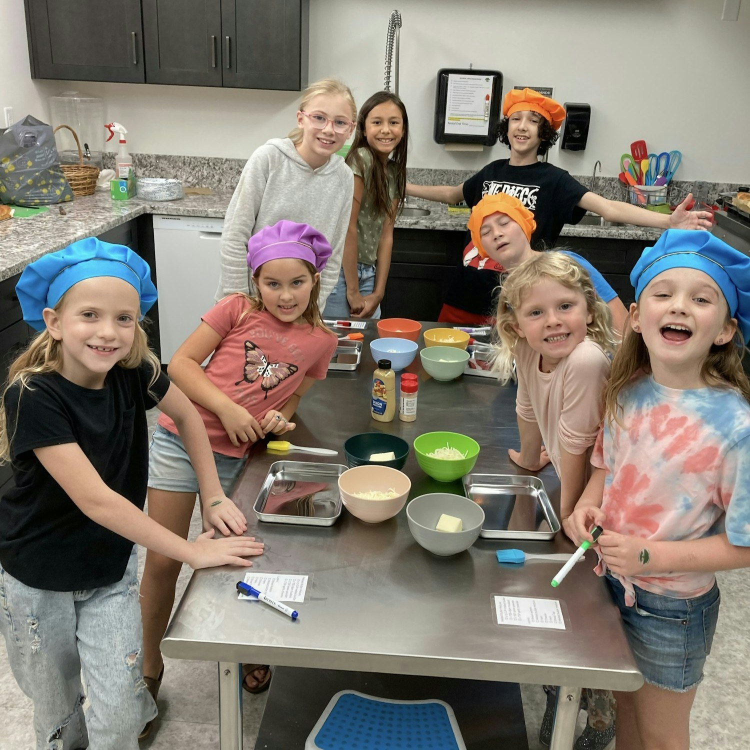 A group of children in colorful chef hats are gathered around a kitchen counter with bowls and cooking ingredients, smiling and engaging.
