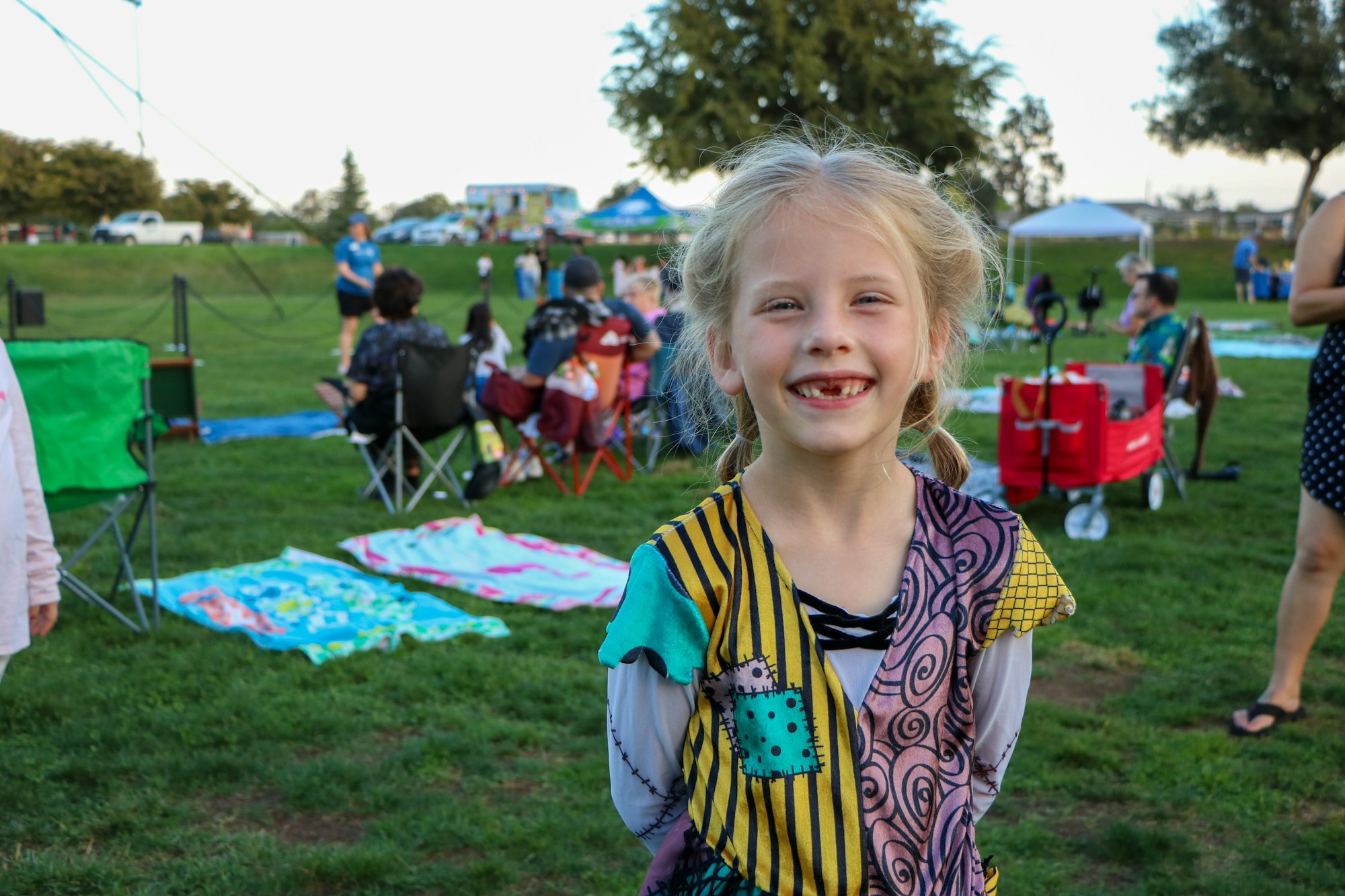 A smiling child with missing teeth at an outdoor event with people, chairs, and a wagon in the background.