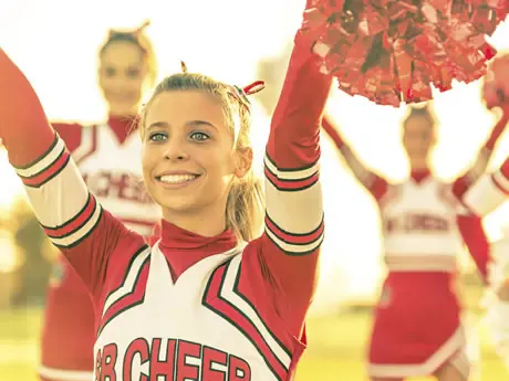 A cheerful cheerleader in a red and white uniform is performing with pom-poms, with teammates in the background.