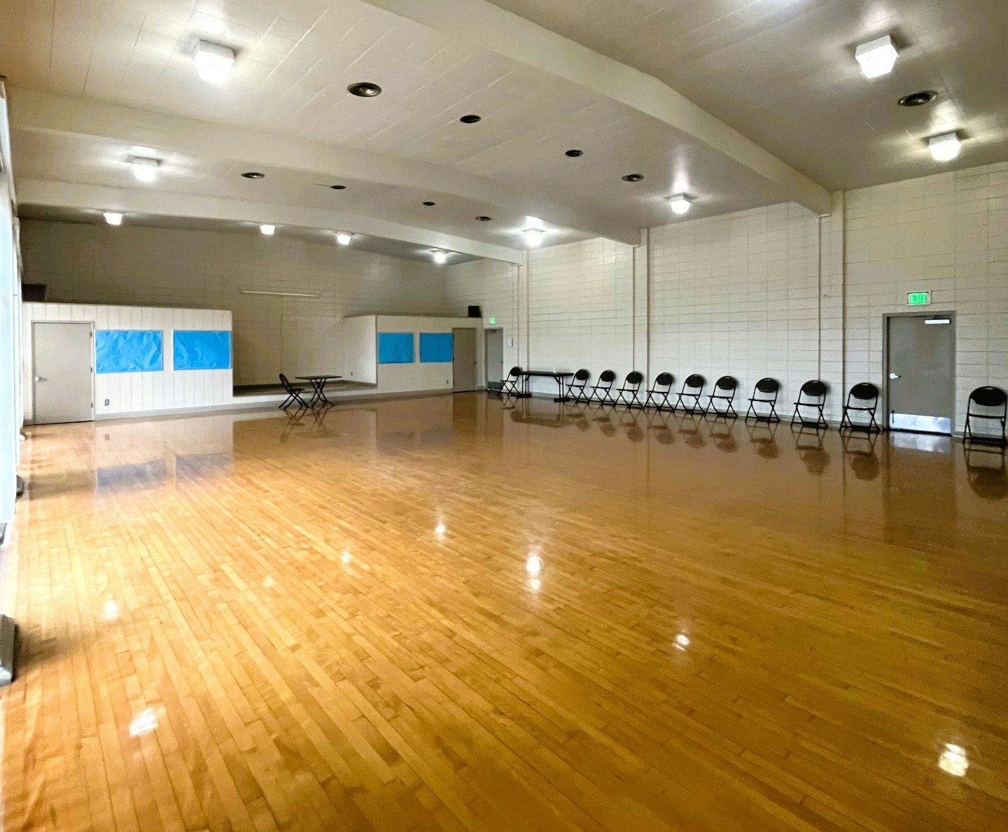 An empty hall with shiny wooden floor, chairs lined up against a wall, tables folded, and white brick walls.