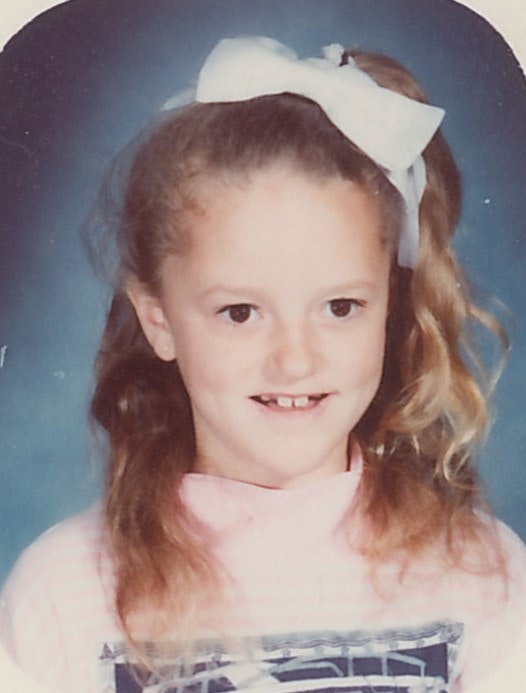 A young child with long hair, a large white bow, and a pink sweater smiling against a blue background.