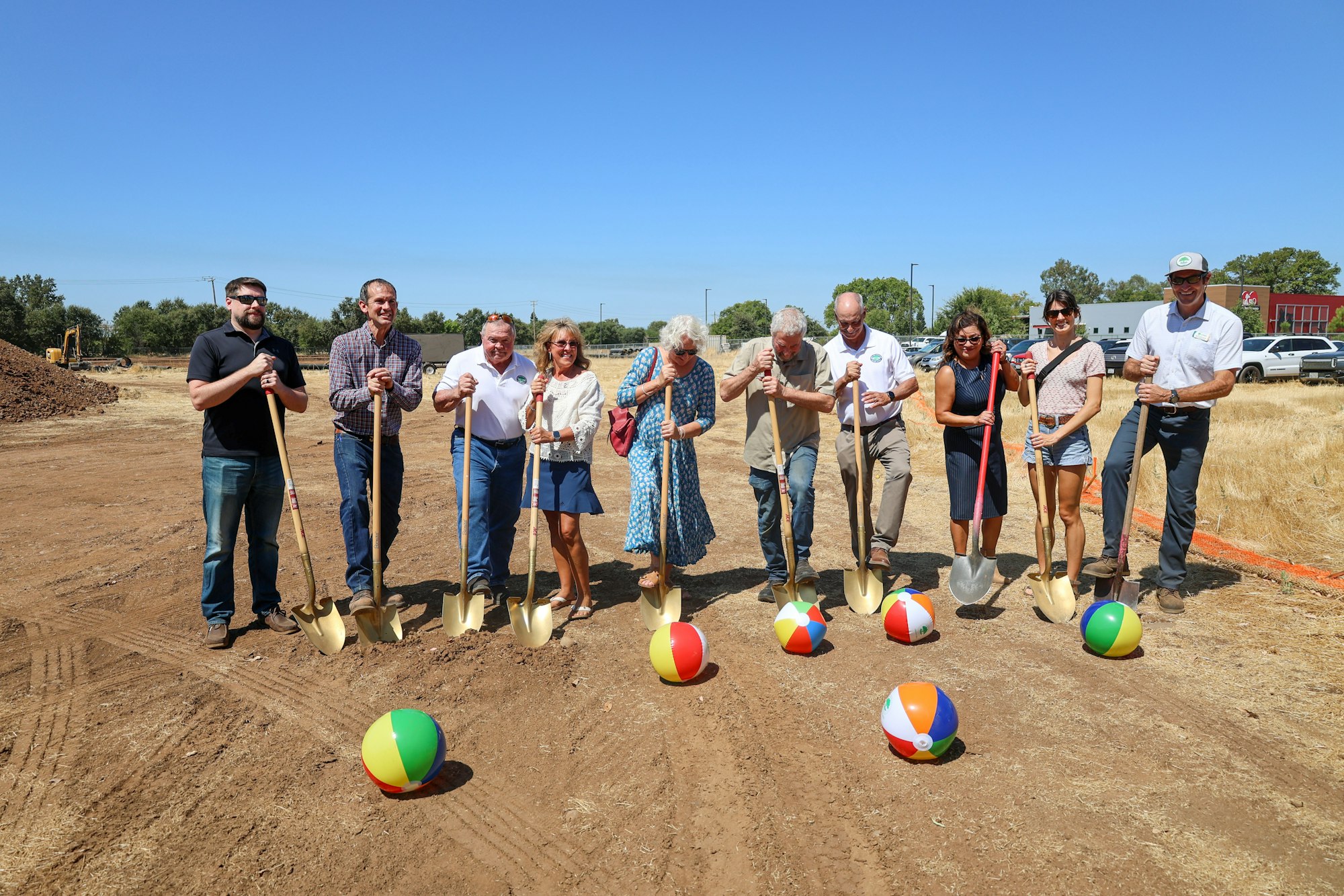 A group of people with shovels stand on a dirt field with colorful beach balls in front of them, likely at a groundbreaking event.