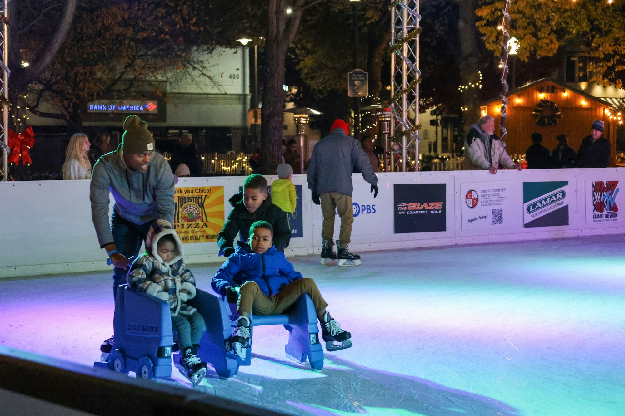 People ice skating; one adult pushing two kids seated on skate aids. Others skate and watch in the festive setting.
