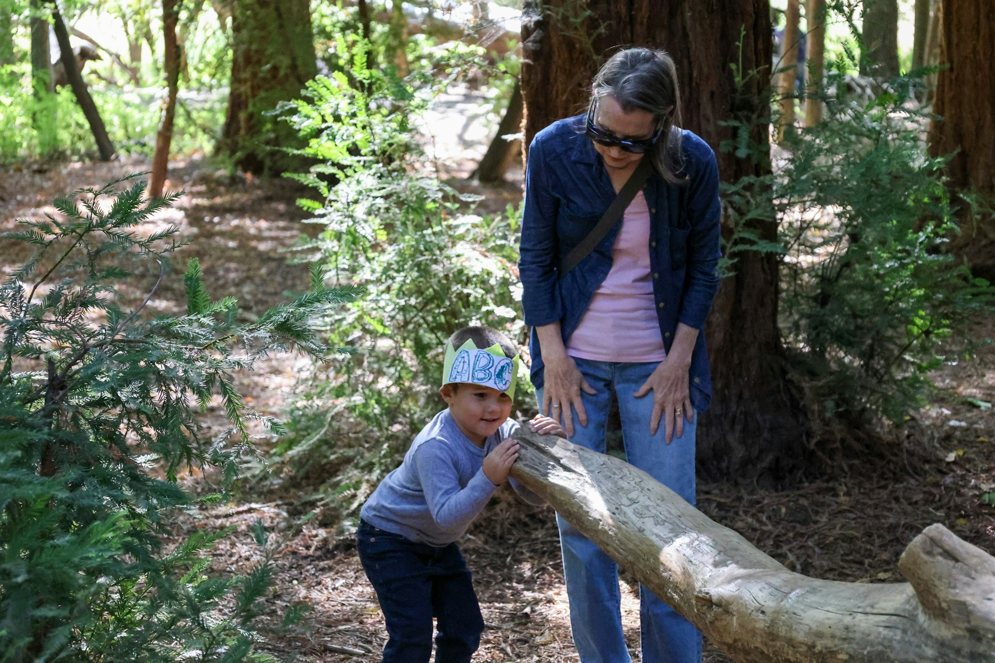 A child in a crown plays with a log while an adult observes in a wooded setting.
