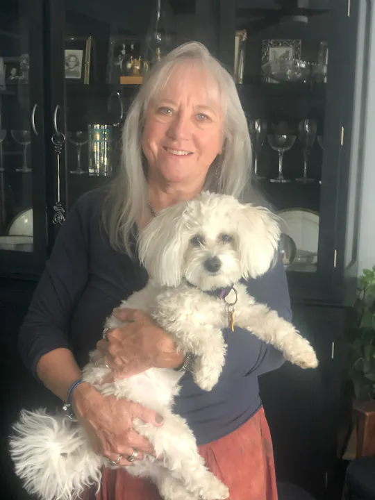 A smiling woman holds a small white dog while standing in front of a dark cabinet with various items displayed.
