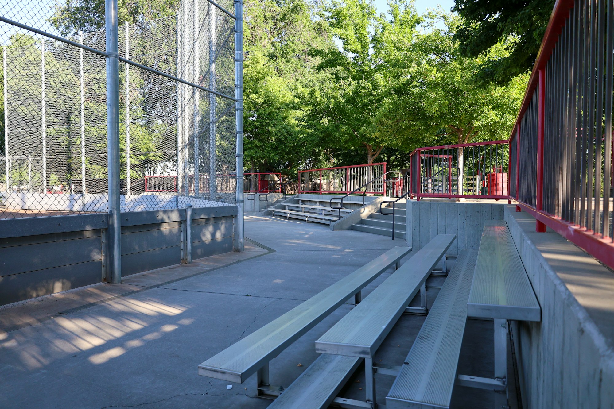 Empty bleachers at an outdoor sports court with trees and fencing.