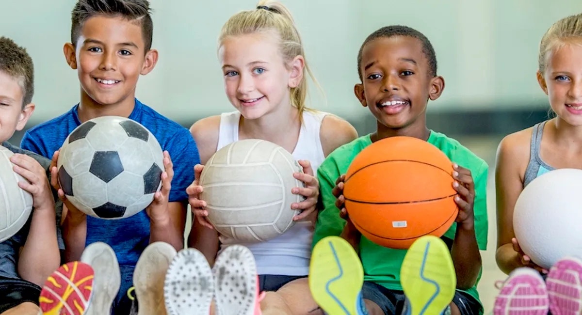 Group of kids with sports balls, smiling, sitting indoors.