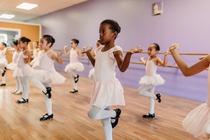 A group of young girls in pink ballet outfits practicing dance in a studio with a purple wall.