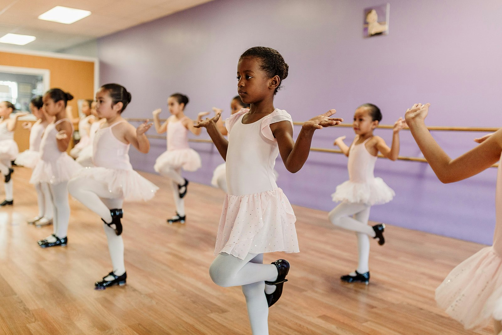 A group of young girls in pink ballet outfits practicing dance in a studio with a purple wall.