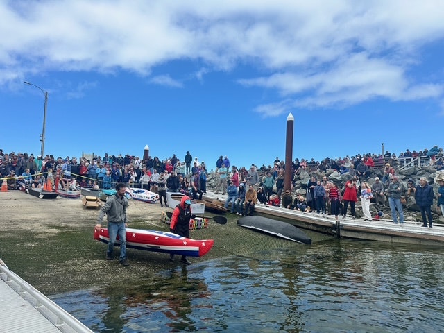 A crowd watches a man carrying a small boat into the water at a lively dockside event.