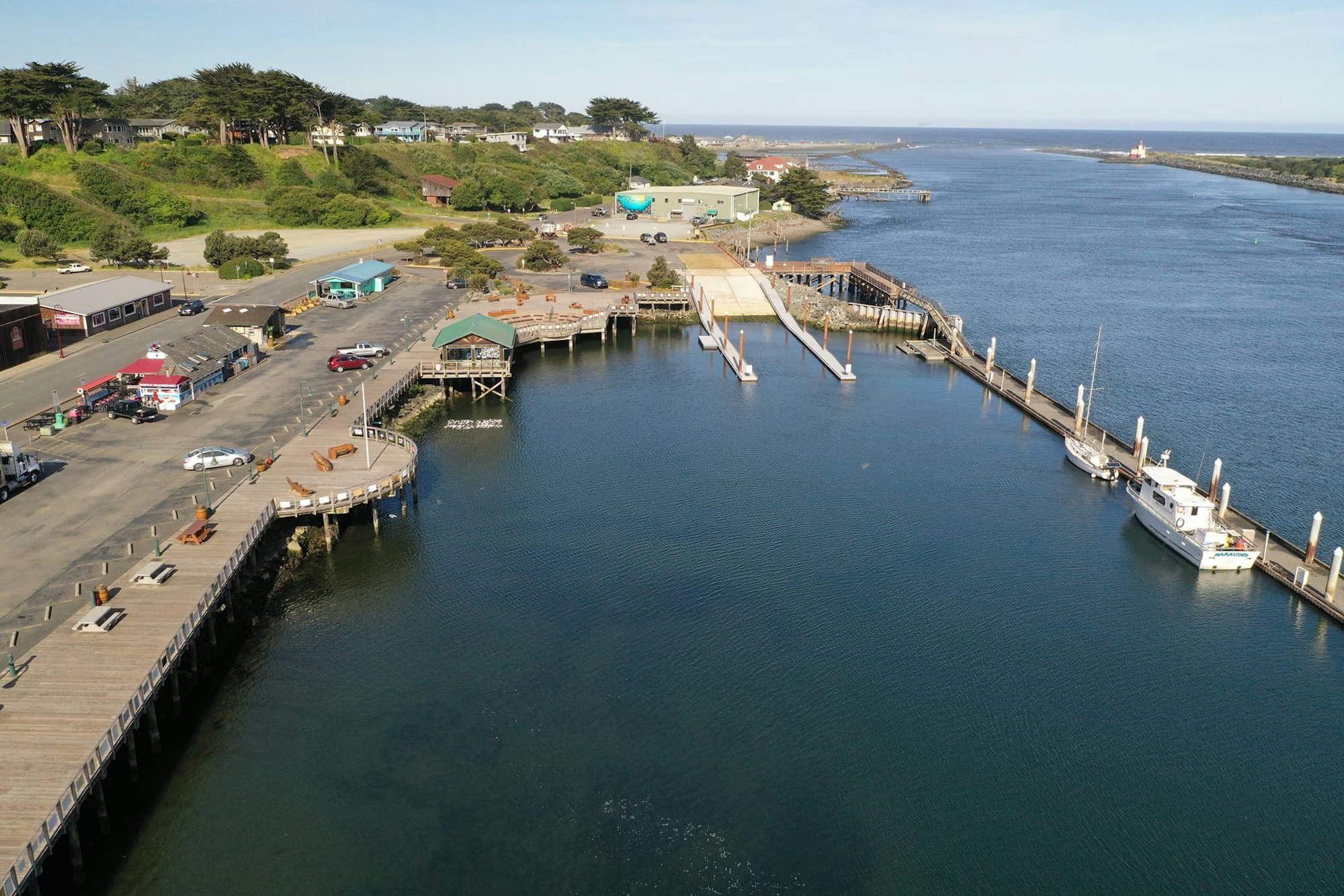 Aerial view of a coastal area with boats, a pier, buildings, and a parking lot near the water.