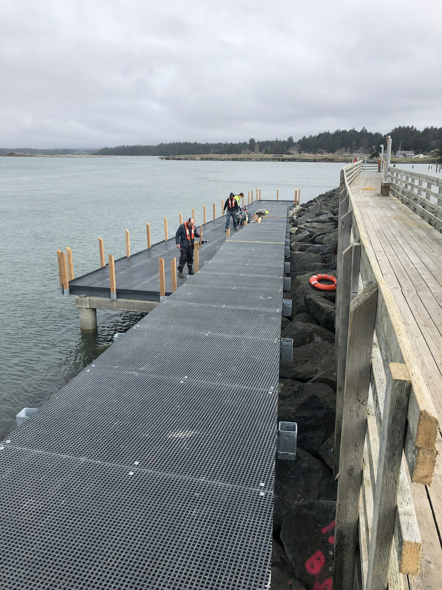 Workers are constructing a metal dock by the water, with a wooden path beside it and rocky shoreline. It's cloudy.