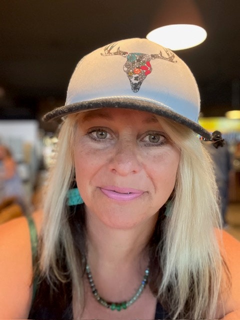 A person wearing a cap with a decorated bull design, earrings, and a necklace, smiling at the camera.