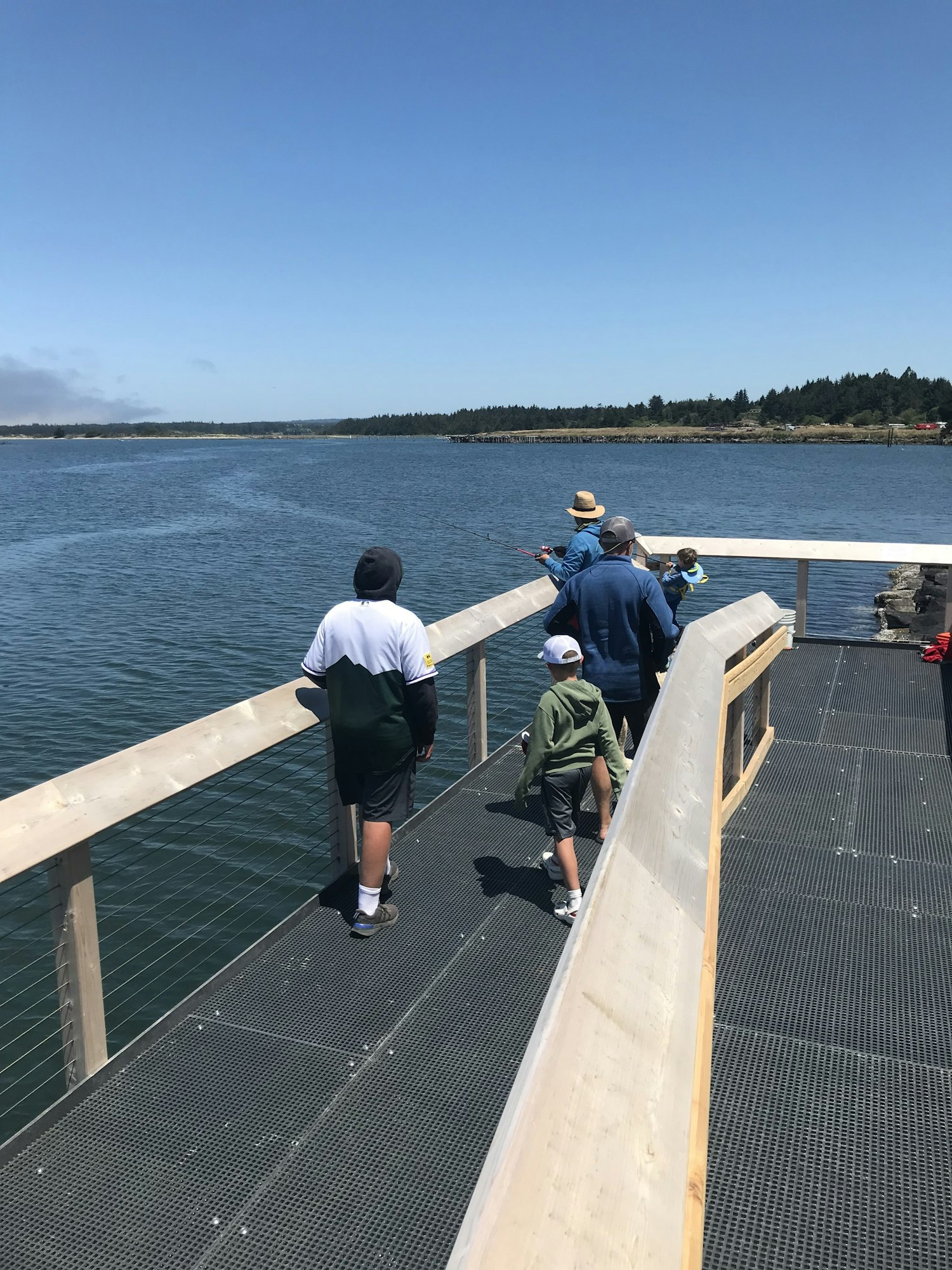A group of people, including children, walking on a pier by the water on a sunny day, with fishing gear visible.