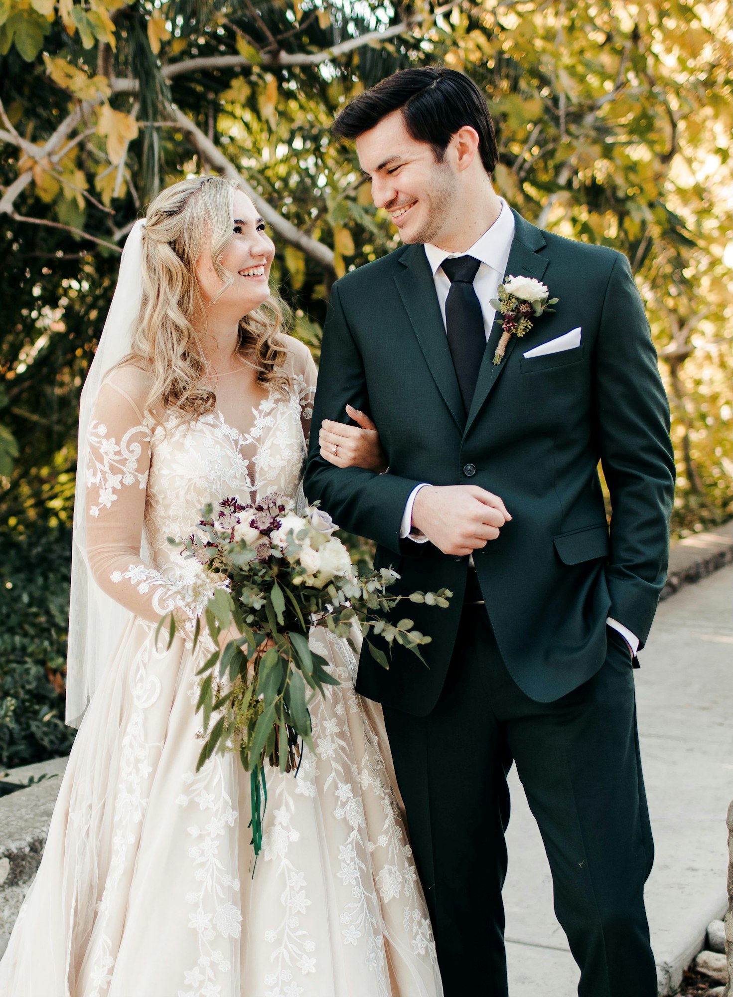 A happy couple in wedding attire, sharing a moment outdoors, with the bride holding a bouquet and surrounded by lush greenery.