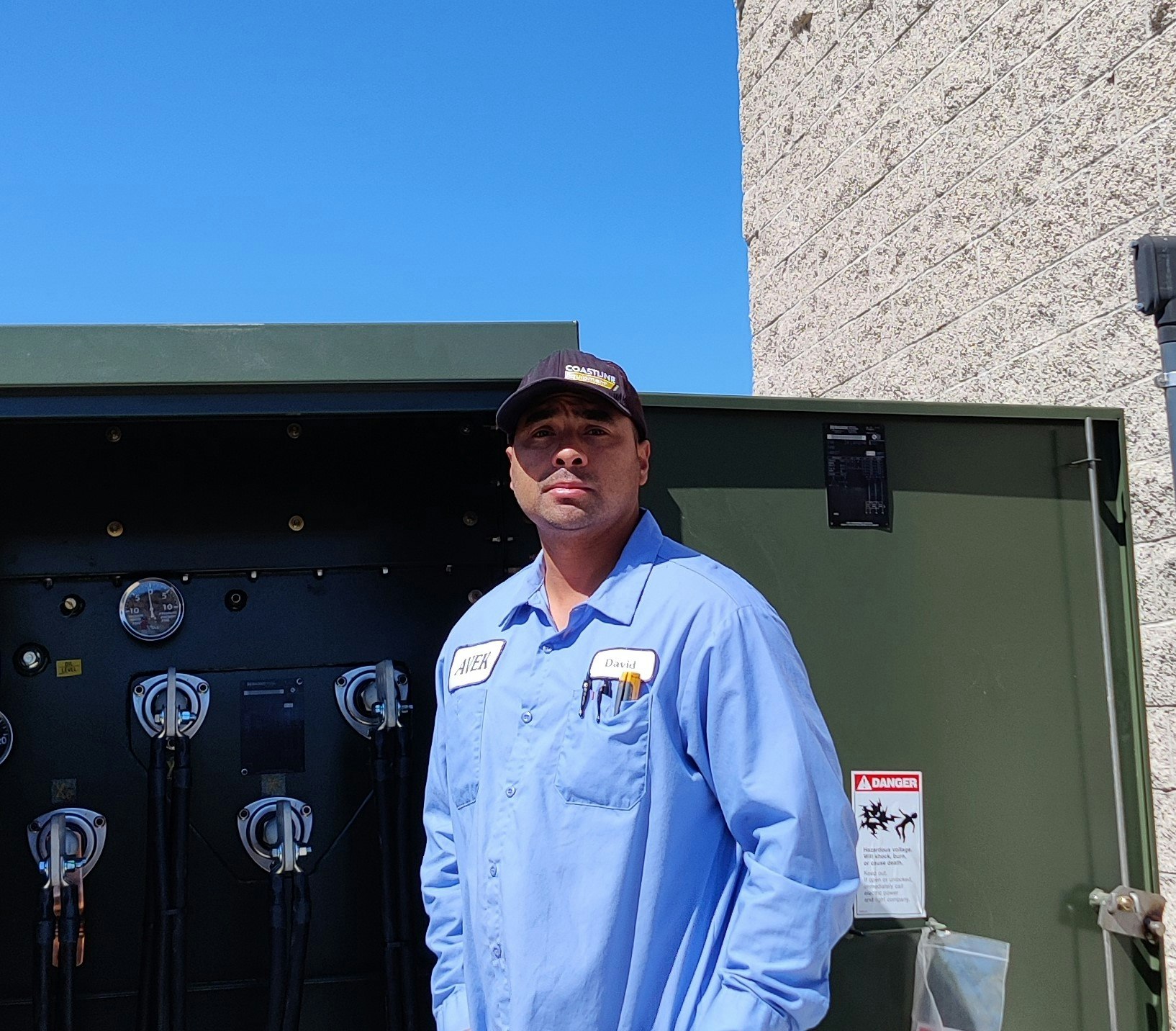 A man in a work uniform stands next to equipment under a clear blue sky, with tools in his pocket and a serious expression.