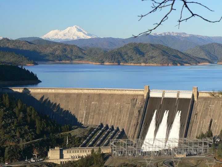 A dam with open spillways, a reservoir, and a snow-capped mountain in the background.
