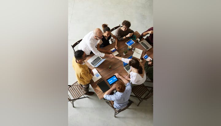 Eight people at a meeting table with laptops, tablets, and notebooks, shaking hands.