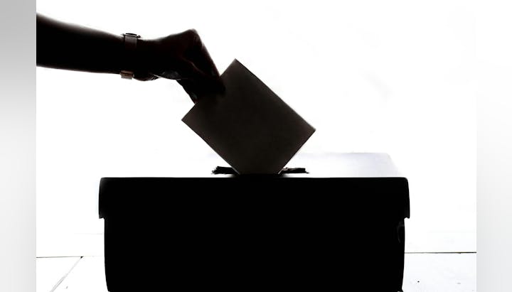 A silhouette of a hand placing a ballot into a voting box.