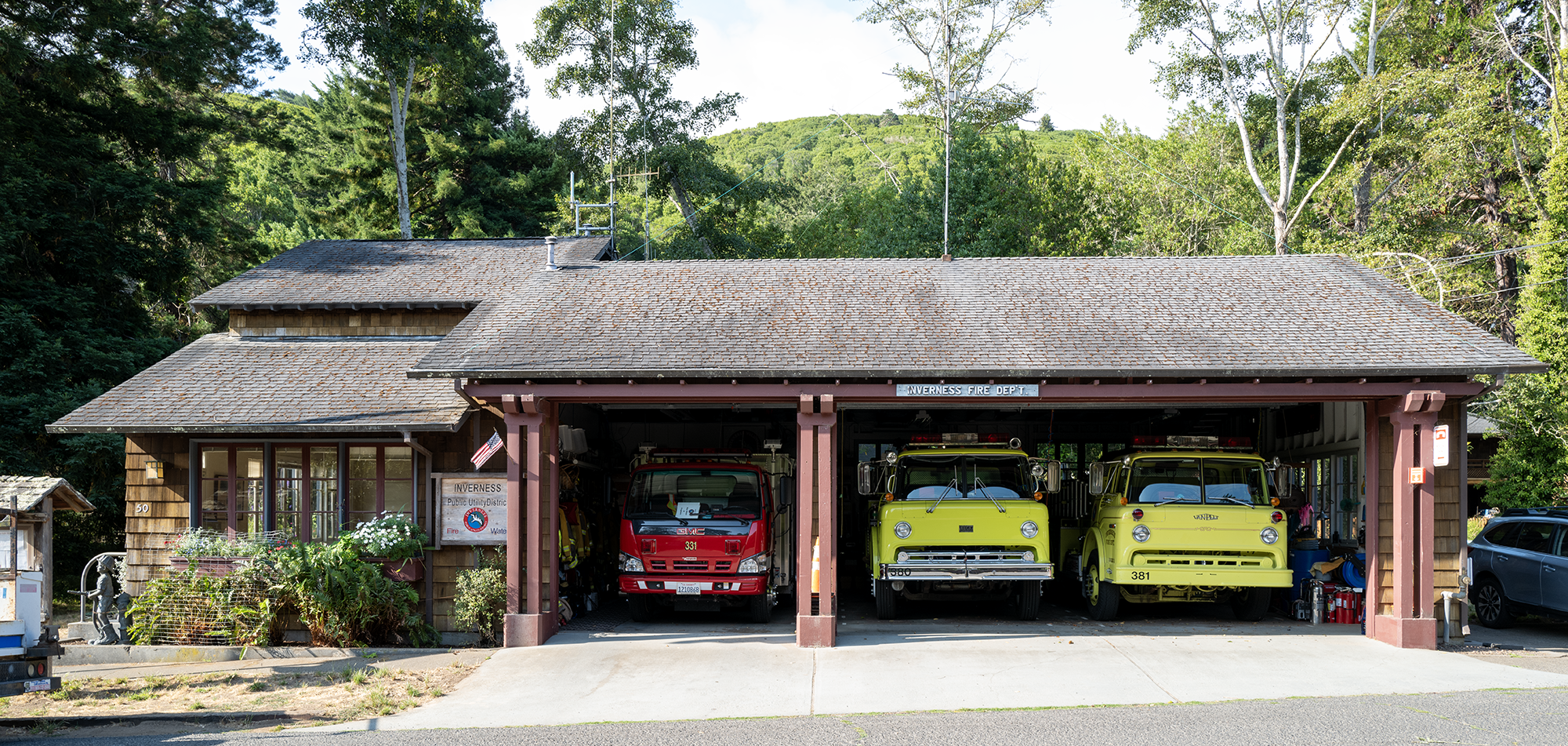 A rustic fire station with red and yellow fire trucks, surrounded by greenery.
