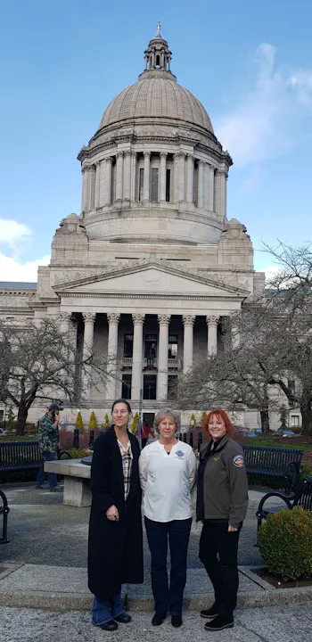 Three women stand in front of a majestic legislative building, with a blue sky and trees in the background.
