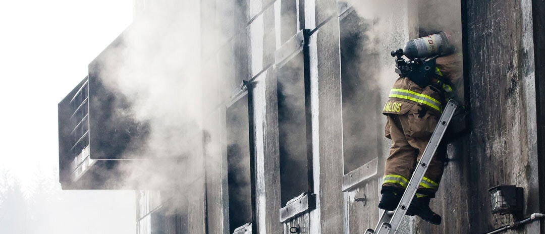 smoke pours out of a burning building with a firefighter on a ladder