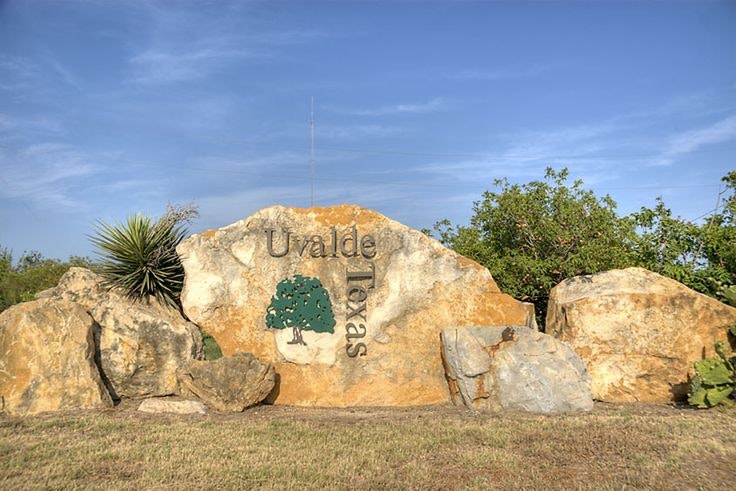 The image shows a large rock sign that reads "Uvalde Texas," surrounded by natural greenery and blue sky.