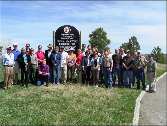 A group of people stand together by a sign for Cherry Creek Valley Ecological Park in a grassy area.