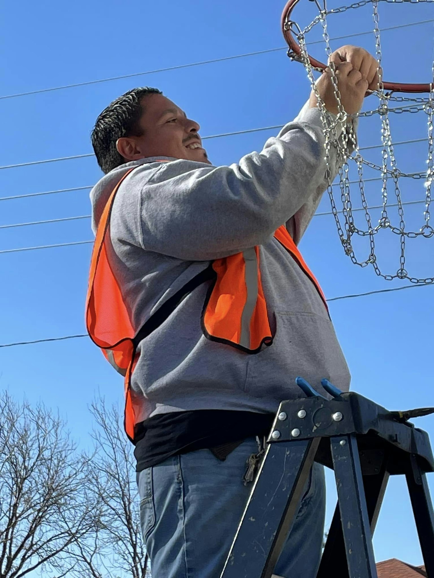 A man in an orange vest on a ladder, fixing a chain basketball net.