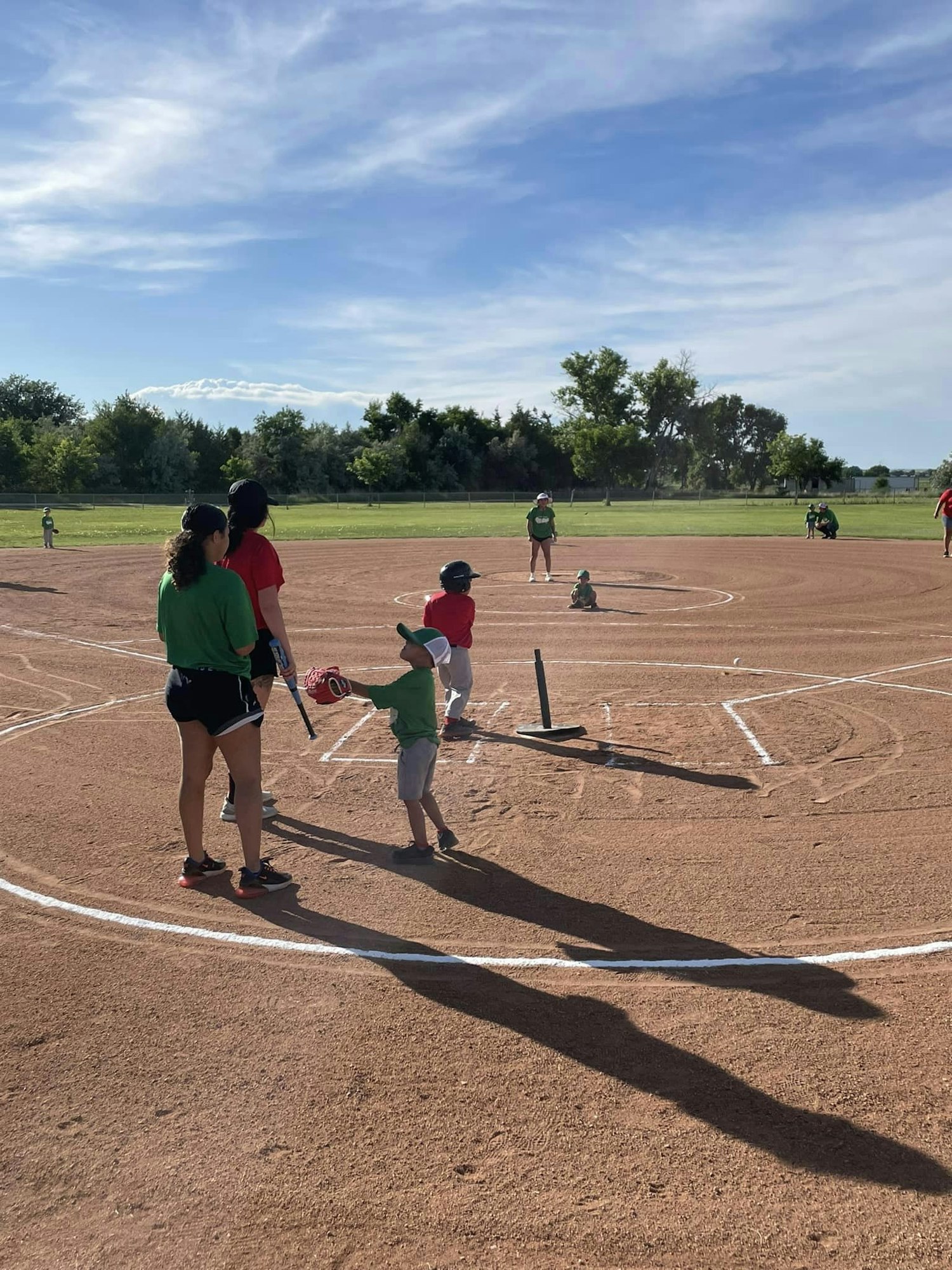 Children playing baseball on a sunny field with adults supervising.
