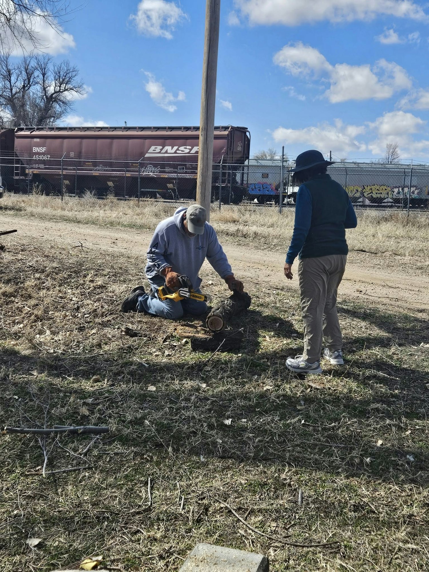 Two people working outdoors with a log and power tool; train car visible in the background.
