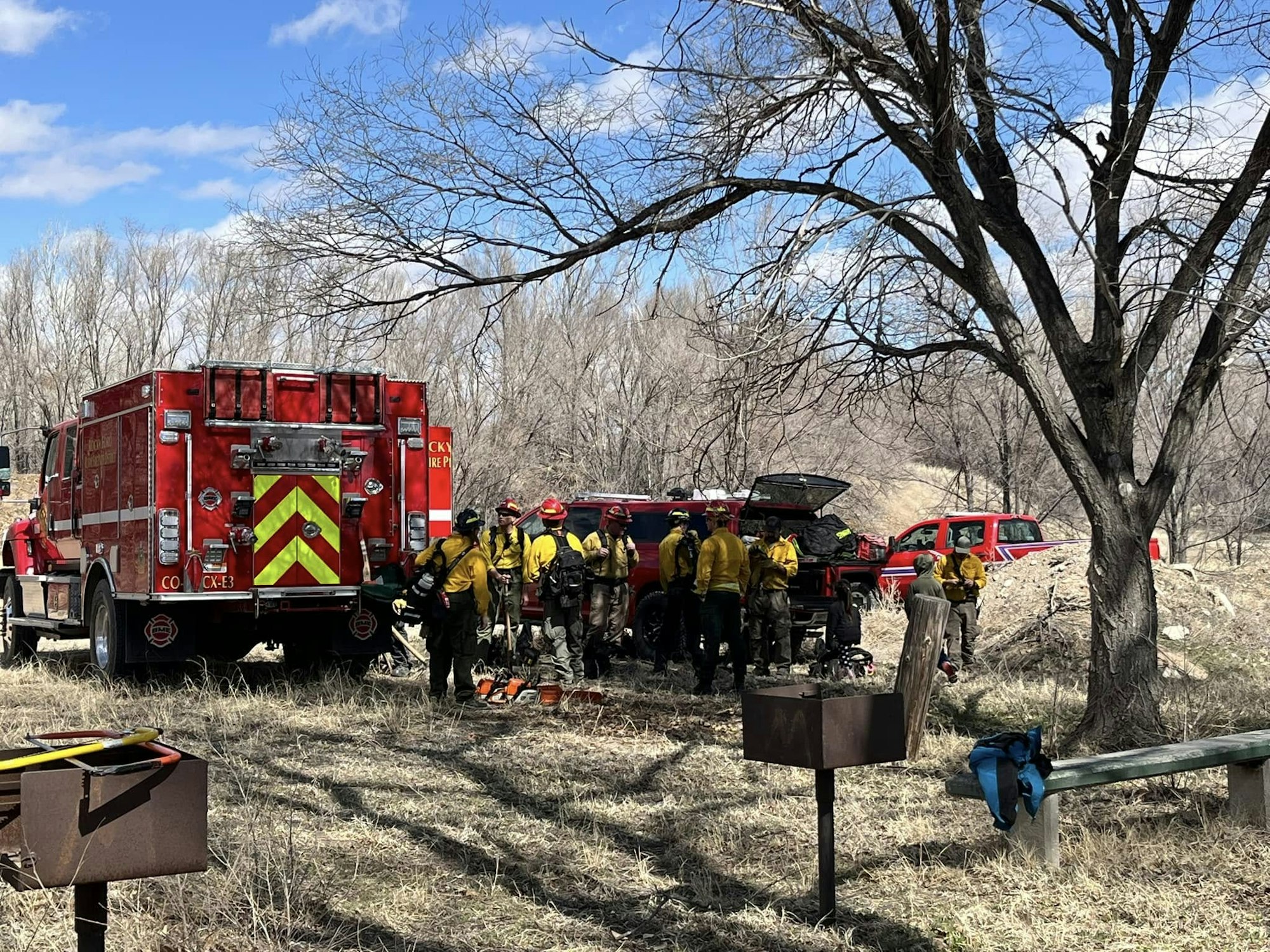 Firefighters in yellow gear stand near red fire trucks and equipment in a wooded area.