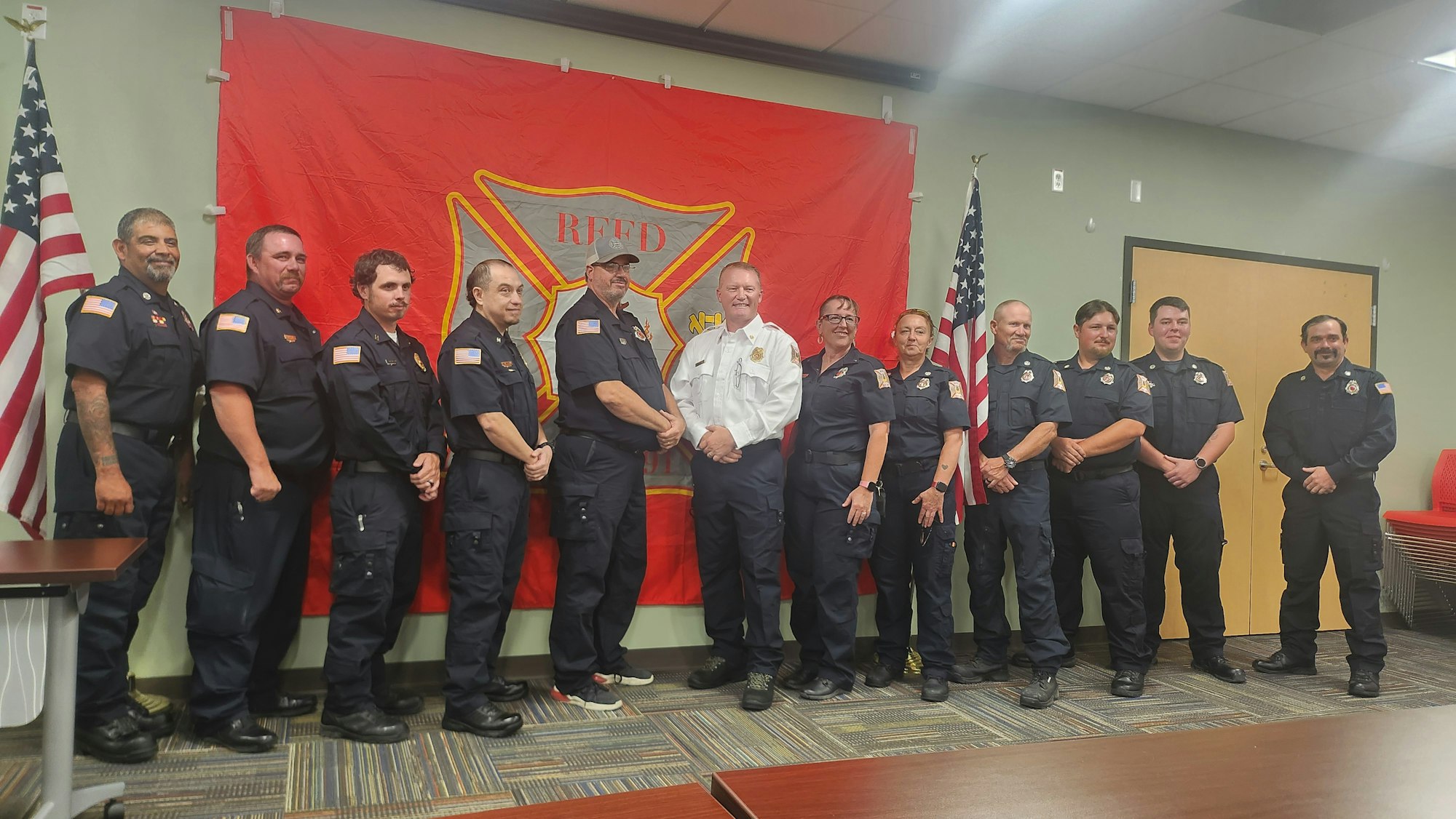 Group of firefighters in uniform in front of a flag with an emblem.