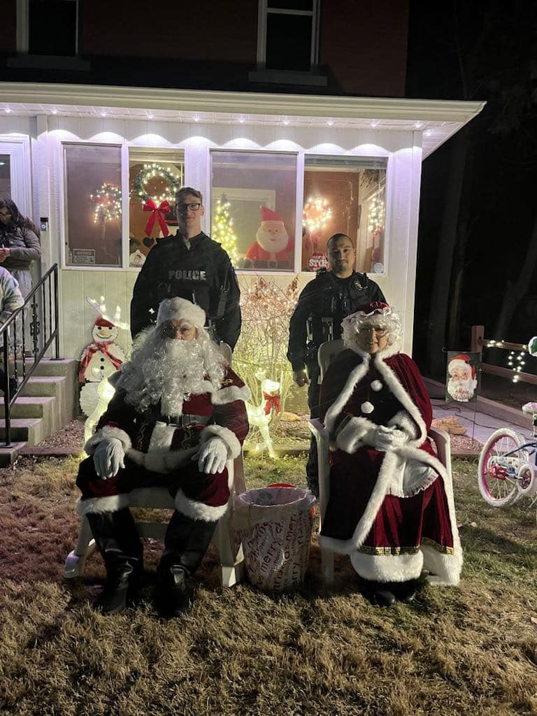 Santa and Mrs. Claus sit on chairs with two police officers standing behind them in front of a festive house.